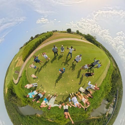 The 360 camera recreates the landscape to look like a perfect, planet-like sphere, with people standing in a grassy circle in the middle of the planet. Blue sunny sky surrounds the "planet".