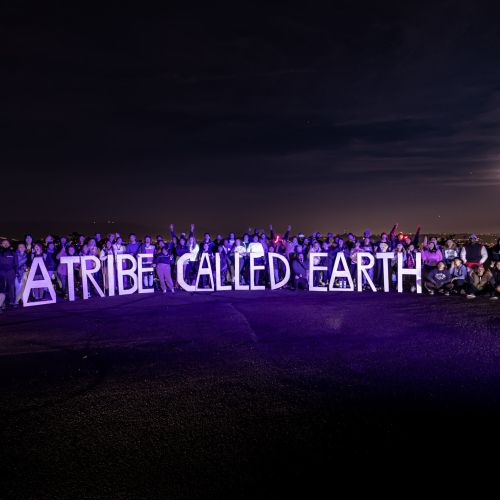 A large group of men and women pose in the dark while holding up letters that spell "A Tribe Called Earth." The people pose against a dark backdrop where city lights can be seen, as well as a bright moon obscured by clouds.