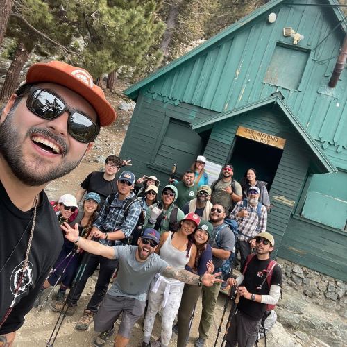 A group of men and women, some with backpacks, hiking poles, and/or hats, pose in front of a teal ski hut on a rocky landscape with coniferous trees.