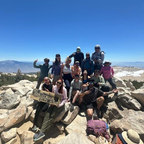 A group of men and women pose on a rocky surface among a mountainous backdrop. They hold a sign that reads "Mt. San Gorgonio elev. 11,503."