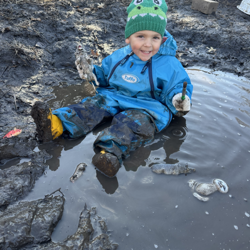 Young child in blue rain suit and green knitted cap sitting in a mud puddle smiling widely.