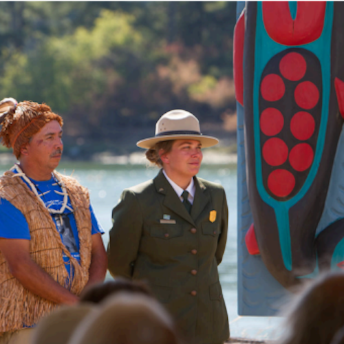 A cultural educator and park service member stand side by side facing a group of people in front of a body of water.