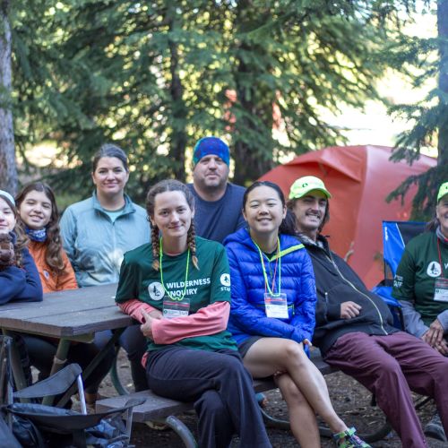 A Military Families Outdoors group smiles while sitting on a picnic bench in Yellowstone National Park. Surrounding them are tall trees, which provide shade for the group.
