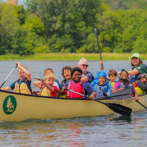 A large group of students smile while paddling their canoe during a Canoemobile program in the Twin Cities. Behind them is lush greenery that lines the edge of the lake.