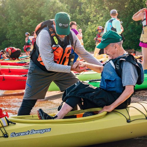 A Wilderness Inquiry staff member helps a participant with cerebral palsy settle into a kayak. Several other participants are also getting into their kayaks behind them.