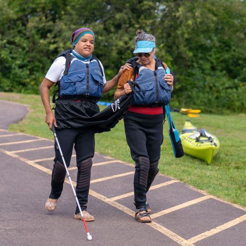 Two participants walk down to the kayak loading area on a Deaf, DeafBlind, and Hard of Hearing affinity trip. One person is using a guide stick and is being guided by another person.
