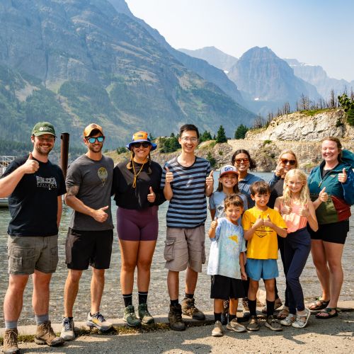 A Military Families Outdoors group poses in front of a picturesque view in Glacier National Park. A lake stretches behind them, with a line of large mountains in the distance.