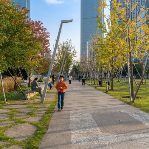 People walk along a broad paved walkway with trees and light posts on both sides and tall buildings in the background.