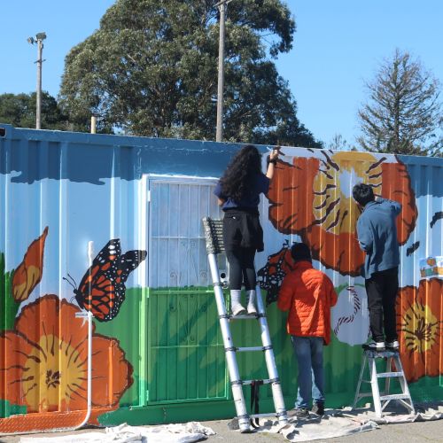 Under a bright blue sky, three youth stand on ladders painting a mural on the side of a shipping container. The mural has orange flowers and monarch butterflies amid a green and blue background.