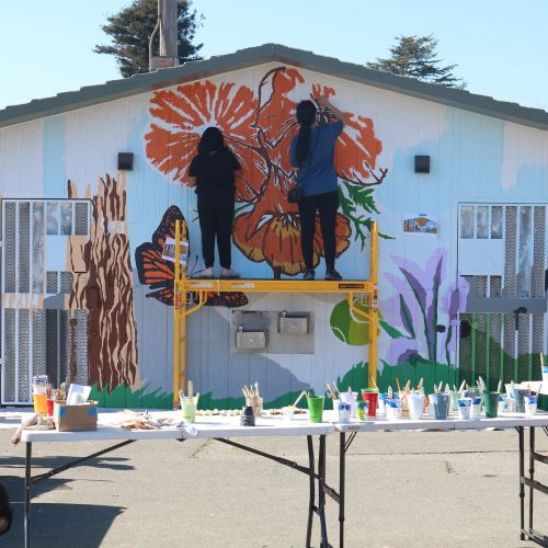 Youth stand on ladders painting a large mural on a building. You can see the beginnings of trees, flowers, and butterflies in the mural. Tables with lots of paint colors are in the foreground. 