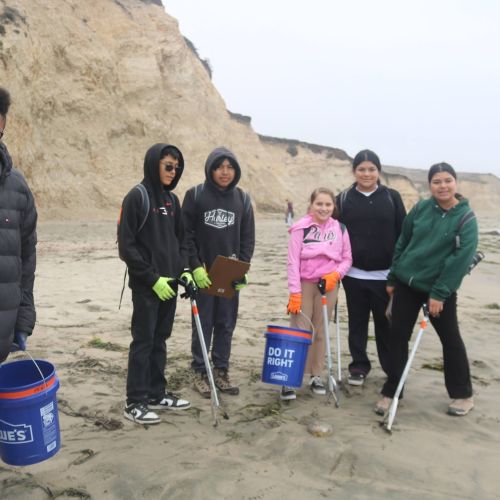 Youth wearing jackets and sweatshirts stand on a sandy beach with blue 5 gallon buckets and trash picking tools. Tall bluffs are behind them. 