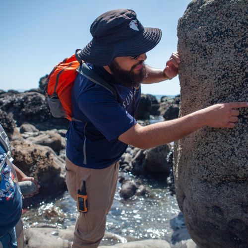 An adult with a hat and orange backpack is pointing to a spot on a large rock while another person with a backpack looks. They're on rocky part of the shoreline with pools of water between rocks visible behind them. 