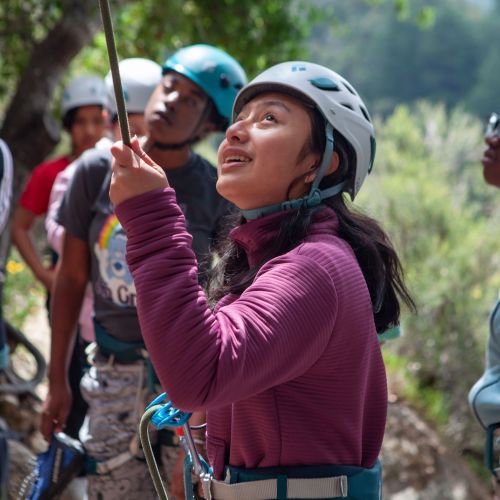 Youth outside wear rock climbing gear. One is gazing upward and holding a rope.