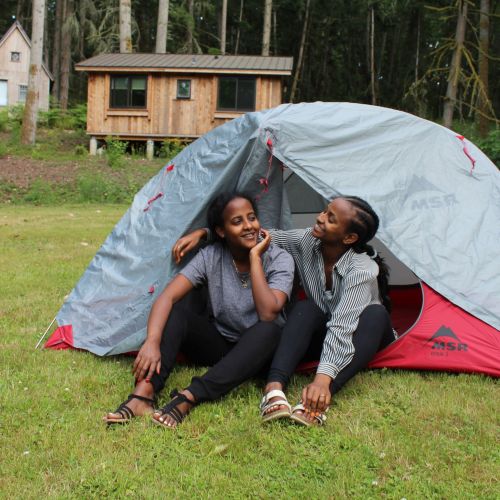 Two participants sit together in a tent and enjoy the outdoors. Their camp is set up in a grassy clearing with thick woods behind them.