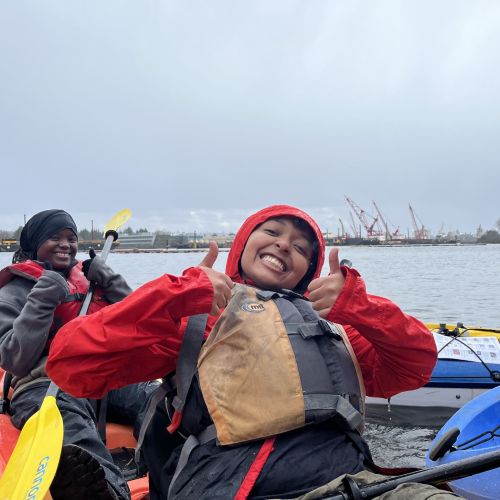  On a cloudy day, two participants give a thumbs-up and wide smiles from where they sit in a kayak together.