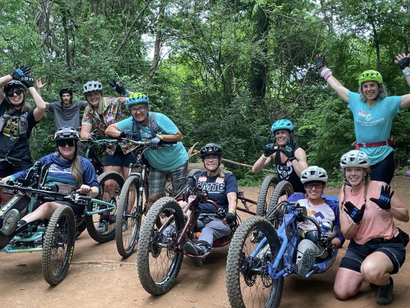 A group of adaptive mountain bike participants stop for a photo with a team of volunteers on bikes behind them, during the AMBC Fall Fest in Knoxville, TN. The adaptive riders are seated on their bikes smiling. Several volunteers are leaning against their bikes on the dirt bike path in front of leafy green trees. 