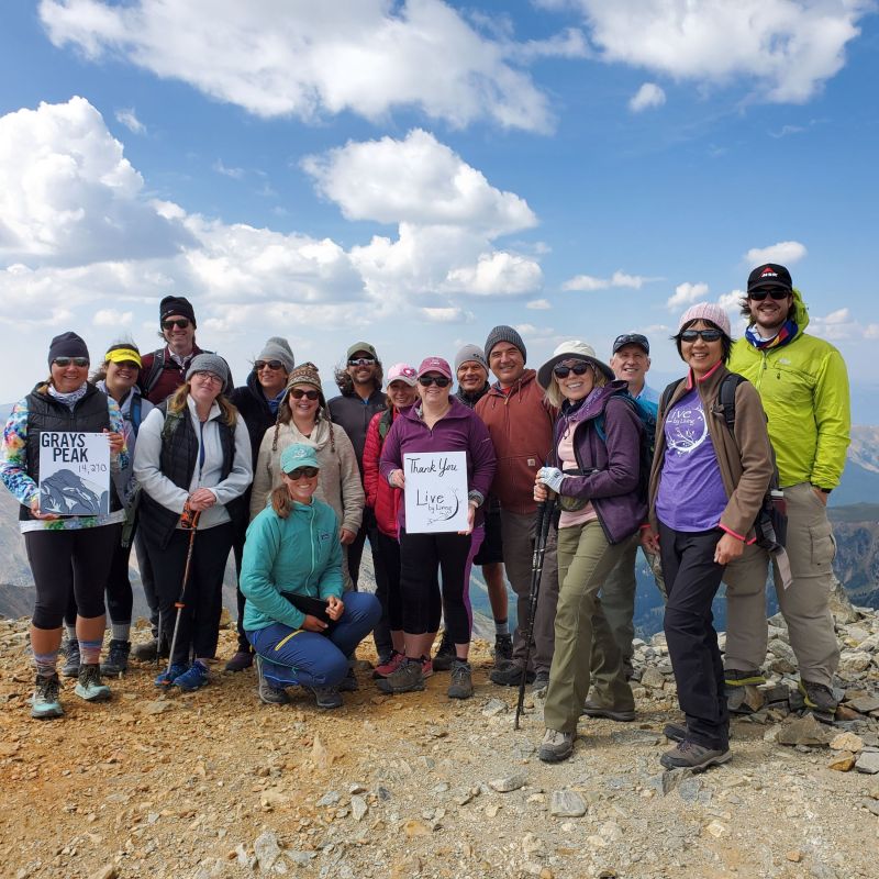 A group of CA to 14k participants stand together at the top of Grays Peak, holding up signs to celebrate their hike’s success. A bright blue sky, filled with fluffy clouds, stretches far behind them.