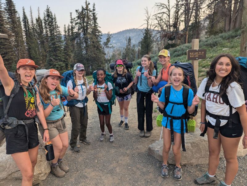 A large group of participants express their excitement at the start of a backpacking trip. Trees and distant mountains fill the horizon behind them.