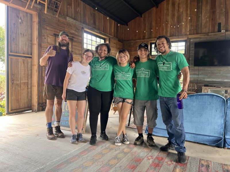 The Program Director Juleon Lewis and Farm Manager Montana Stoval line up for a picture with four Assistant Crew Leaders (ACLs) in a cabin living room. Lewis poses with a thumbs-up and a bright smile, which Stoval and the ACLs also wear on their faces.