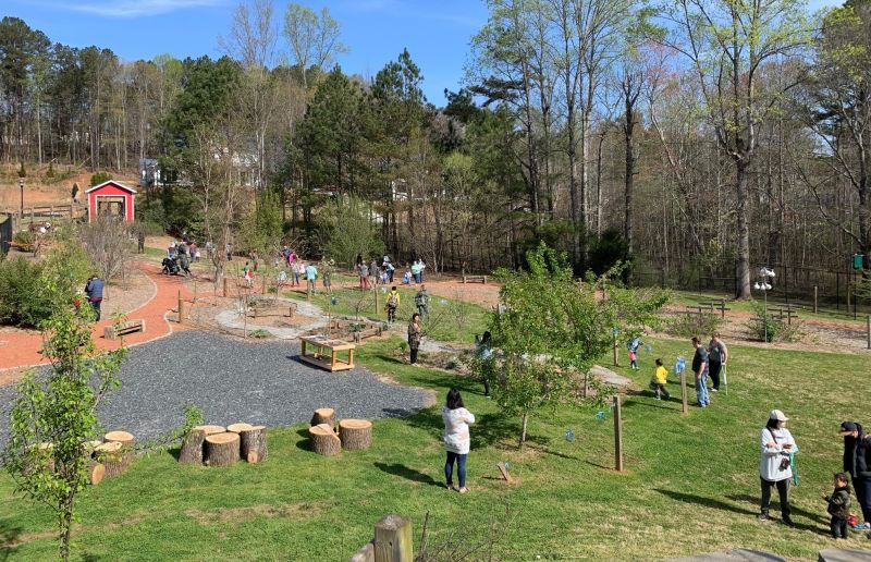 A view from the top of an earthen mound showing playground structures, fruiting plants, and winding pathways in the Orchard. People walk around the park, observing and enjoying nature.