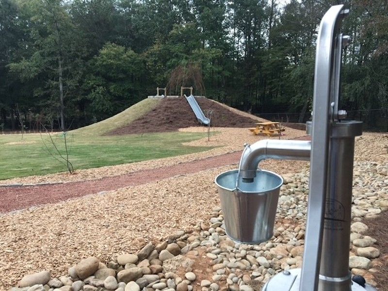 A hand water pump stands in the foreground, with an earthen mound and hillside slide visible in the background.