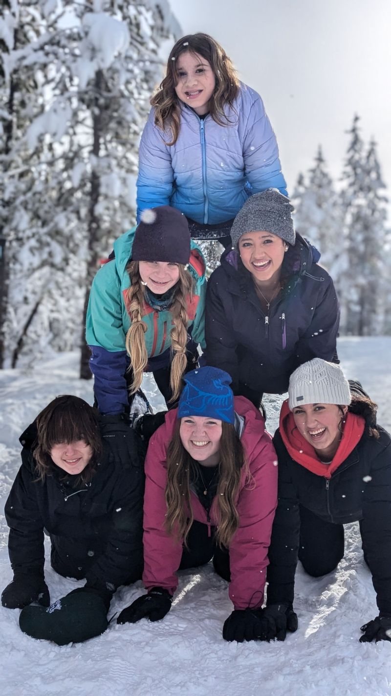 Six beaming girls stack on top of each other to create a human tower in the middle of a snowy, sunny day.