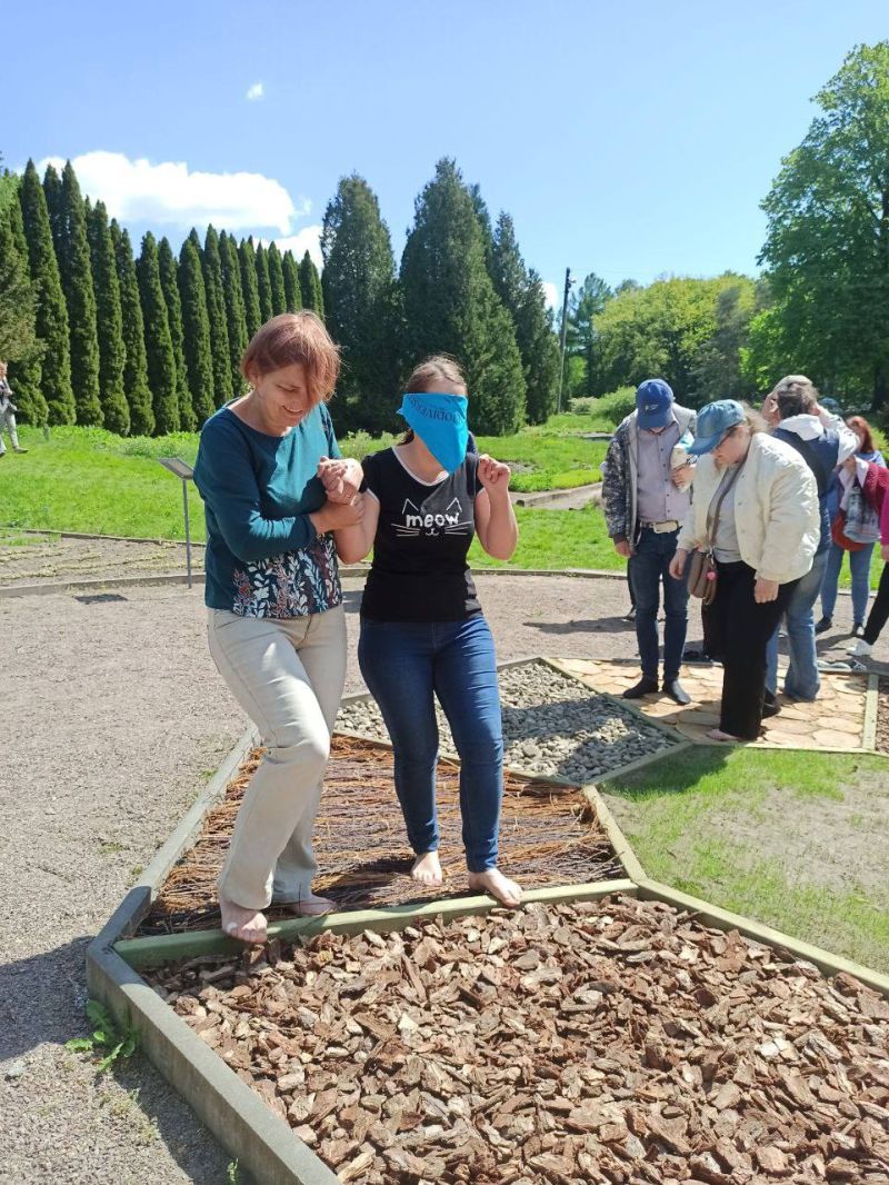 An educator guides a barefoot, blindfolded person by the arm across an outdoor tactile circle—an octagonal path with each segment made of a different natural material for sensory exploration. In the background, a group of people observe and interact with the tactile circle.