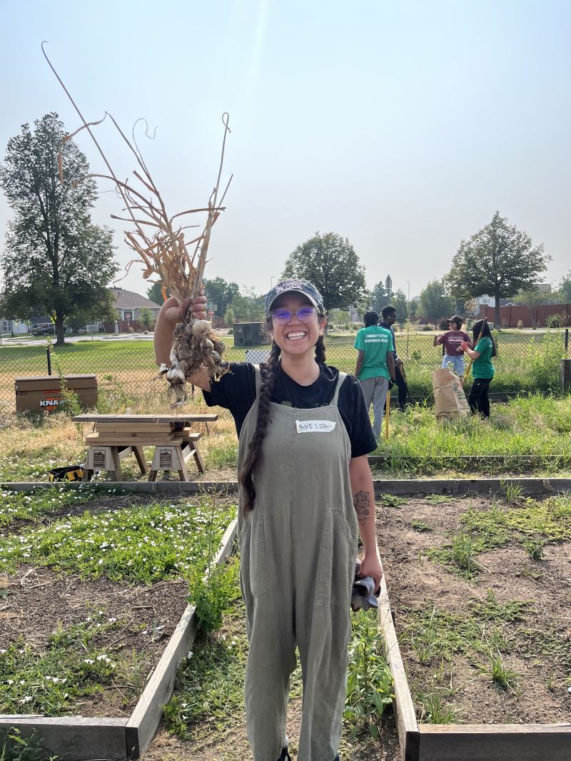 An adult wearing glasses, a blue baseball cap, and taupe colored overalls is smiling and holding a bunch of garlic aloft in their right hand. In the background are raised garden beds and people tending them.