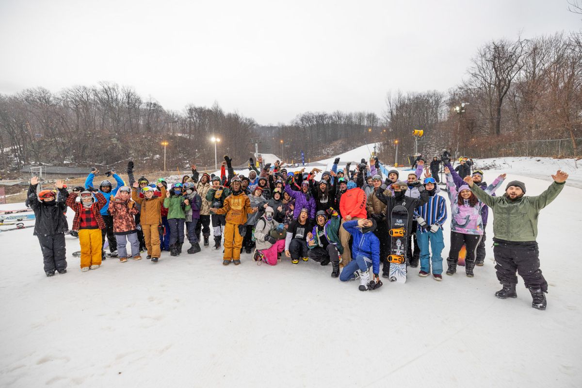 A large group of participants wearing winter gear happily raise their arms and pose for a picture. Around them is a flat, snowy field; barren trees line the horizon behind them.